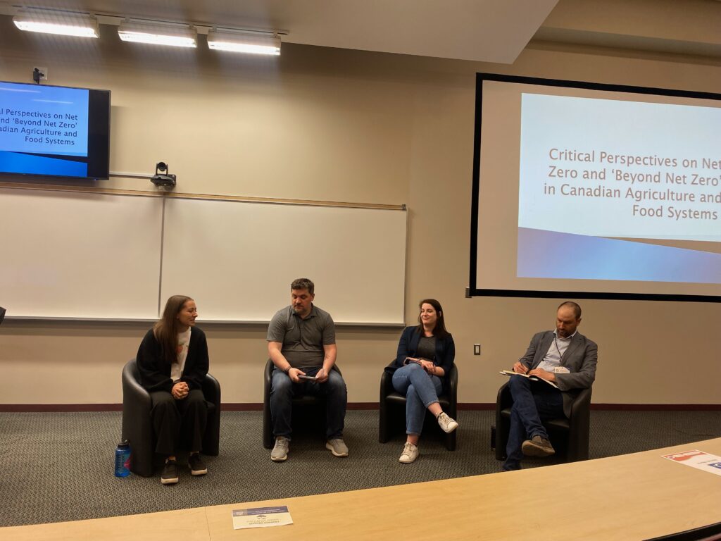 Four adults sit in a row at the front of a classroom. Behind them there is a slideshow with the text "Critical Perspectives on Net Zero and ‘Beyond Net Zero’ in Canadian Agriculture and Food Systems" 